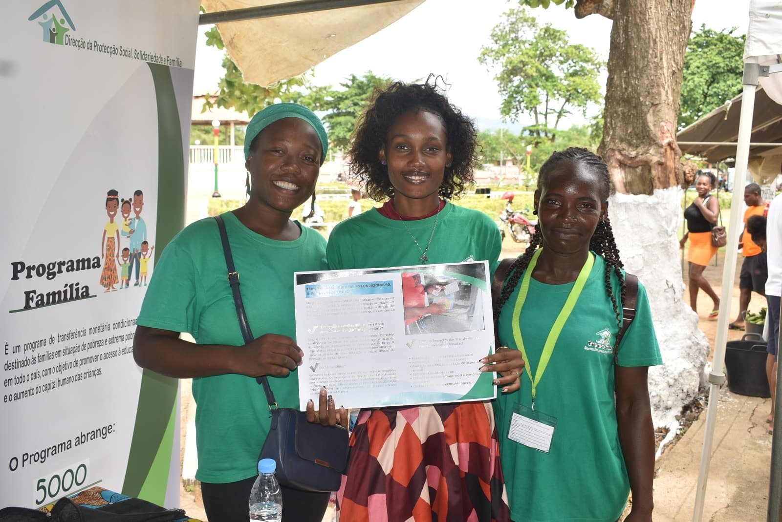 Três mulheres sorridentes em camisetas verdes segurando um cartaz informativo do Programa Família.
